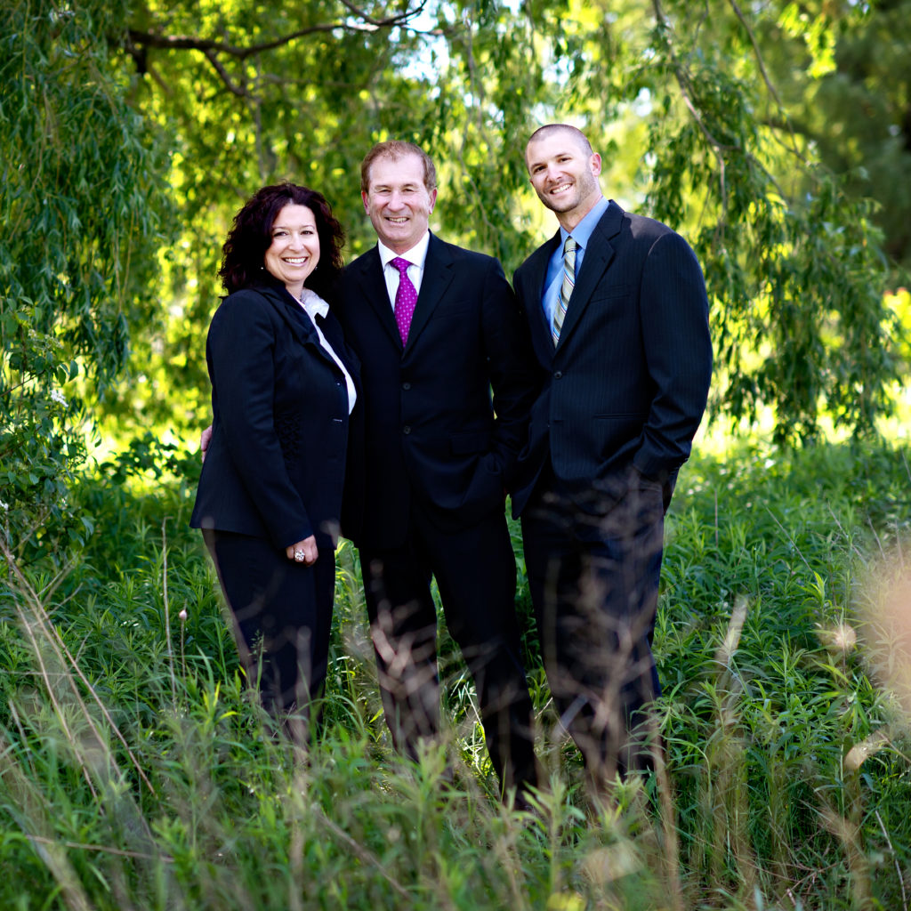 Janet McKenzie, Larry Freiburger, and Scott Freiburger standing in a row and smiling at camera. Tall grass and large green trees blow in the background.