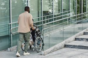 iStock-1415350137 A person pushing a person in a wheelchair up an accessibility ramp in the front of a building.